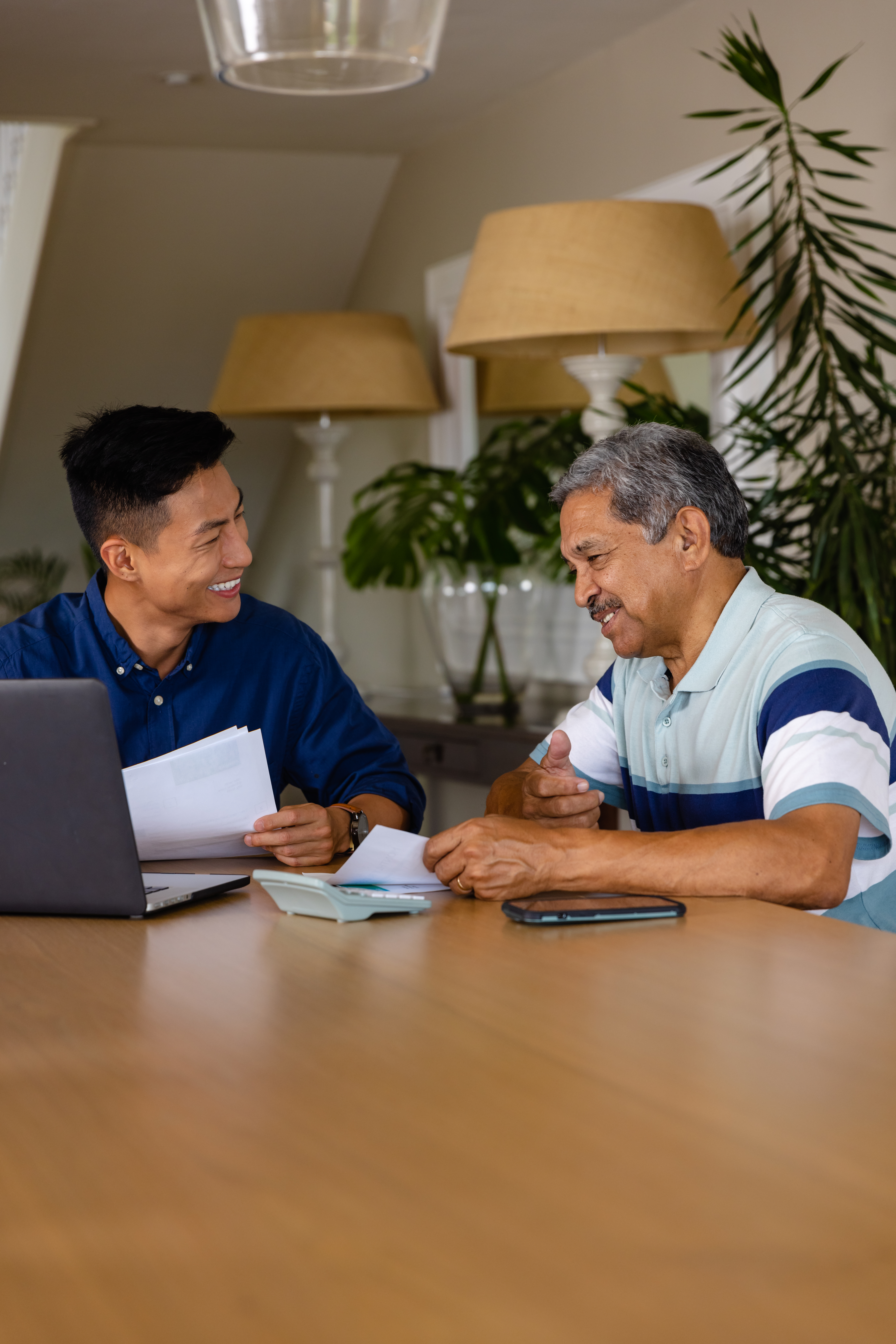 Happy diverse financial advisor and senior man discussing paperwork and using laptop in dining room. Finance, retirement, domestic life, communication and senior lifestyle, unaltered.