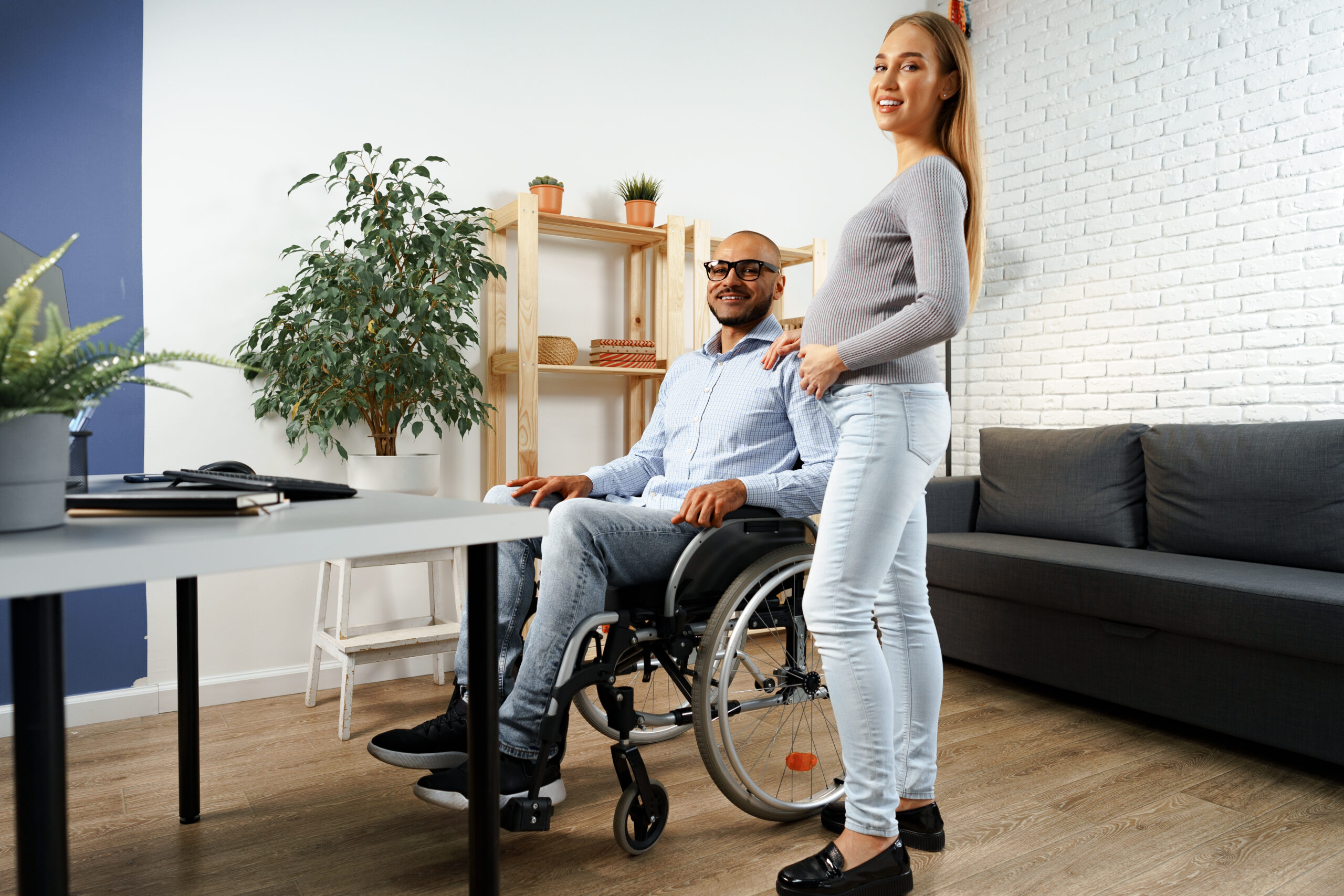 Pregnant woman holding a hand of her disabled husband sitting in a wheelchair, indoors